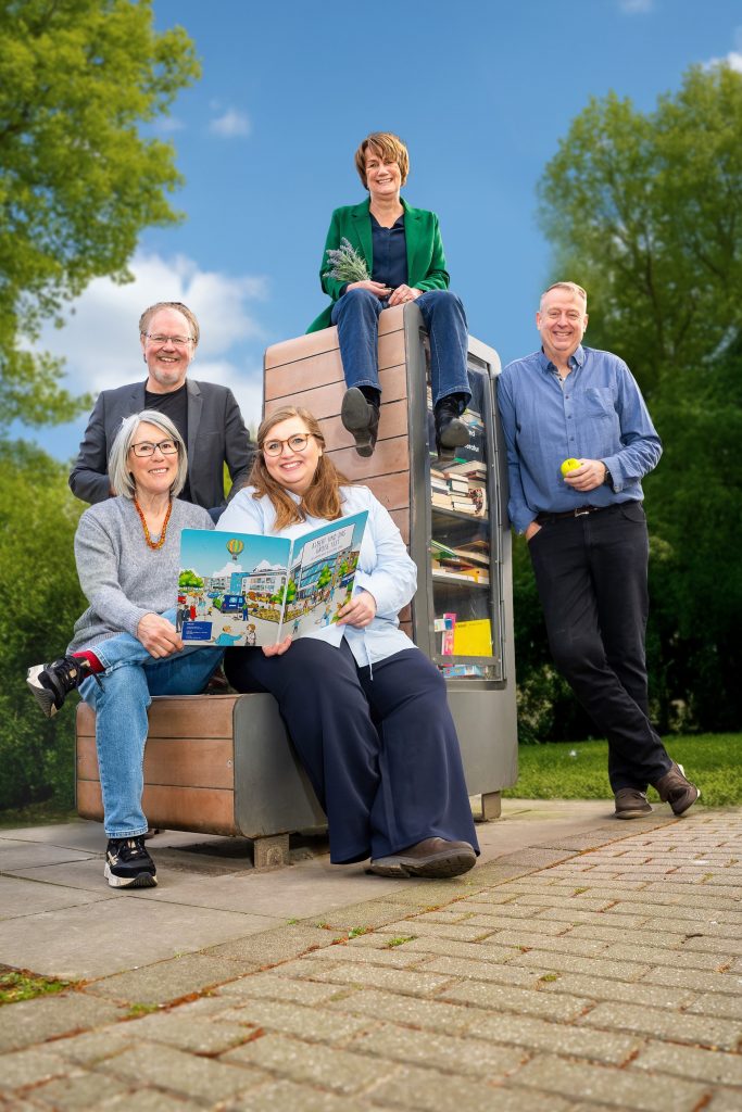 Fünf Personen stehen und sitzen um einen Kiosk im Freien. Eine Frau hält ein Buch mit Bildern von Wohnprojekten. Die Stimmung ist freundlich und entspannt, umgeben von grüner Natur.