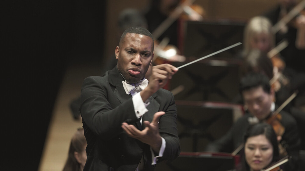 Ein Dirigent in formeller Kleidung führt ein Orchester. Er zeigt Ausdruck und Konzentration, während Musiker im Hintergrund mit ihren Instrumenten spielen. Die Stimmung ist dynamisch und professionell.